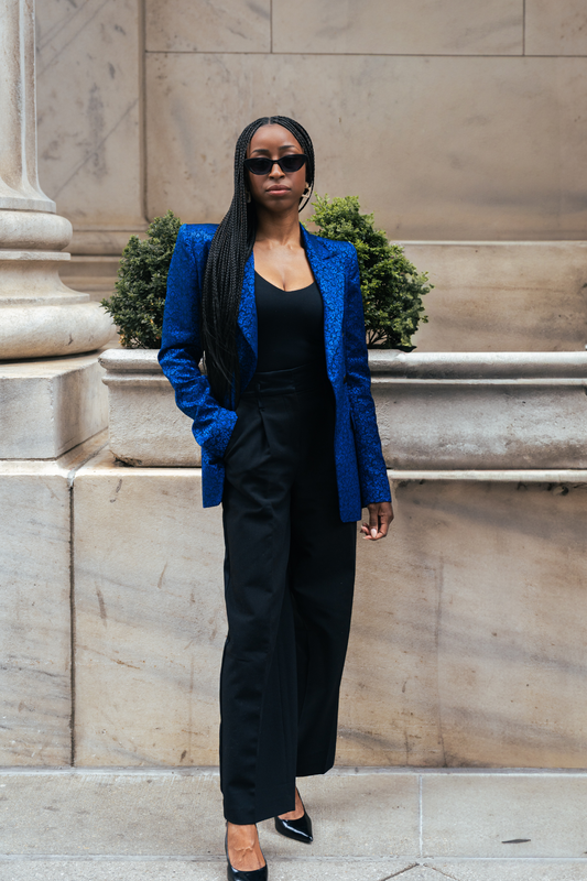Woman in a blue blazer and black outfit standing against a stone wall.