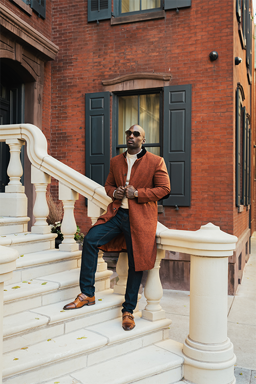 Man in a brown coat sitting on stone steps in front of a brick building