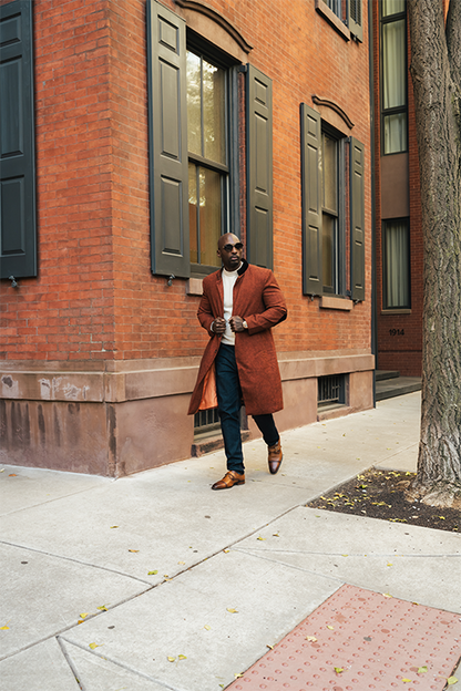 Man in a brown coat standing on a sidewalk next to a brick building
