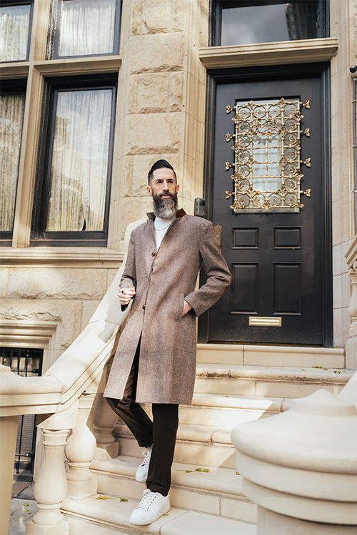 Man in a long coat standing on stone steps in front of a building with a decorative door.