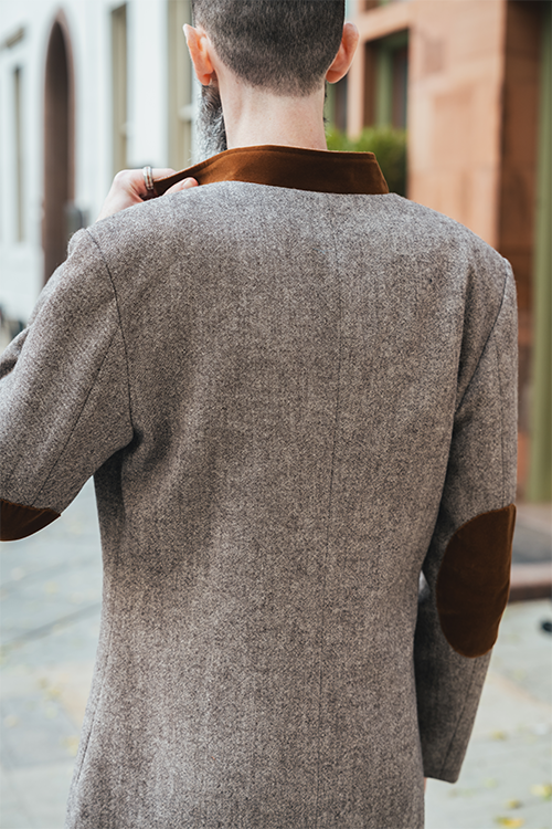 Person wearing a brown coat with brown elbow patches outdoors.