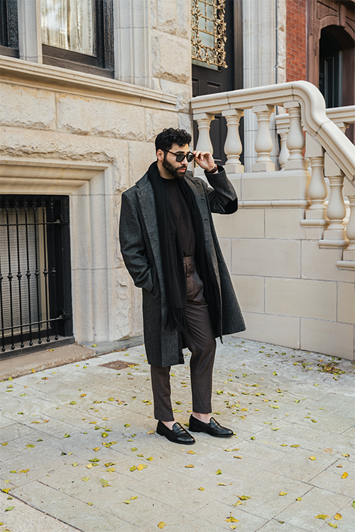 Man in a dark coat and suit standing on a sidewalk with a building in the background