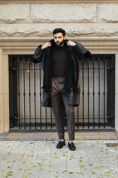 Man wearing a black coat over a black shirt and brown pants, standing in front of a stone building with metal bars.