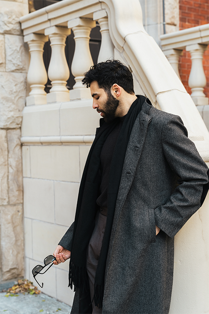Man wearing a long dark coat standing against a stone wall.