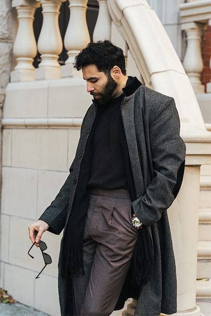 Man wearing a long dark coat and sunglasses, standing against a stone building.