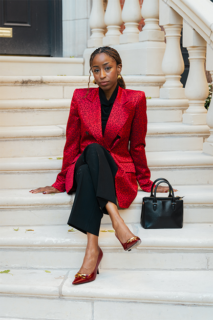 Woman in a red blazer and black pants sitting on white steps with a black handbag.