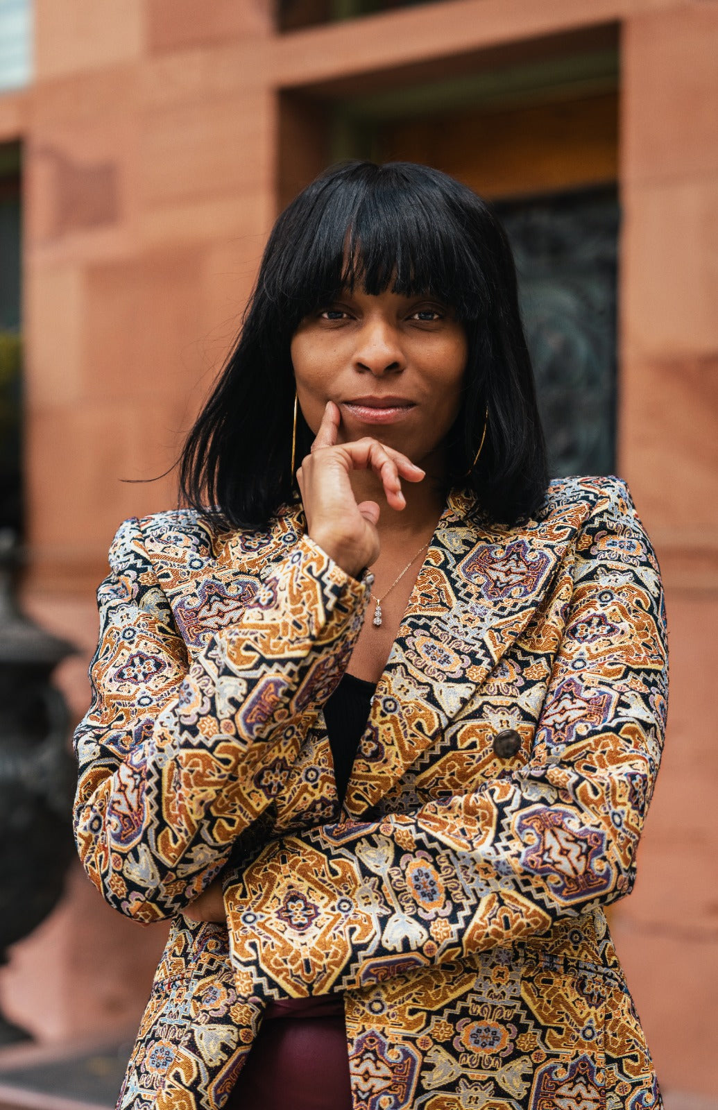 Aria Roadcloud wearing her patterned blazer in gold, brown, and cream tones, posing outdoors against a terracotta-colored building background.